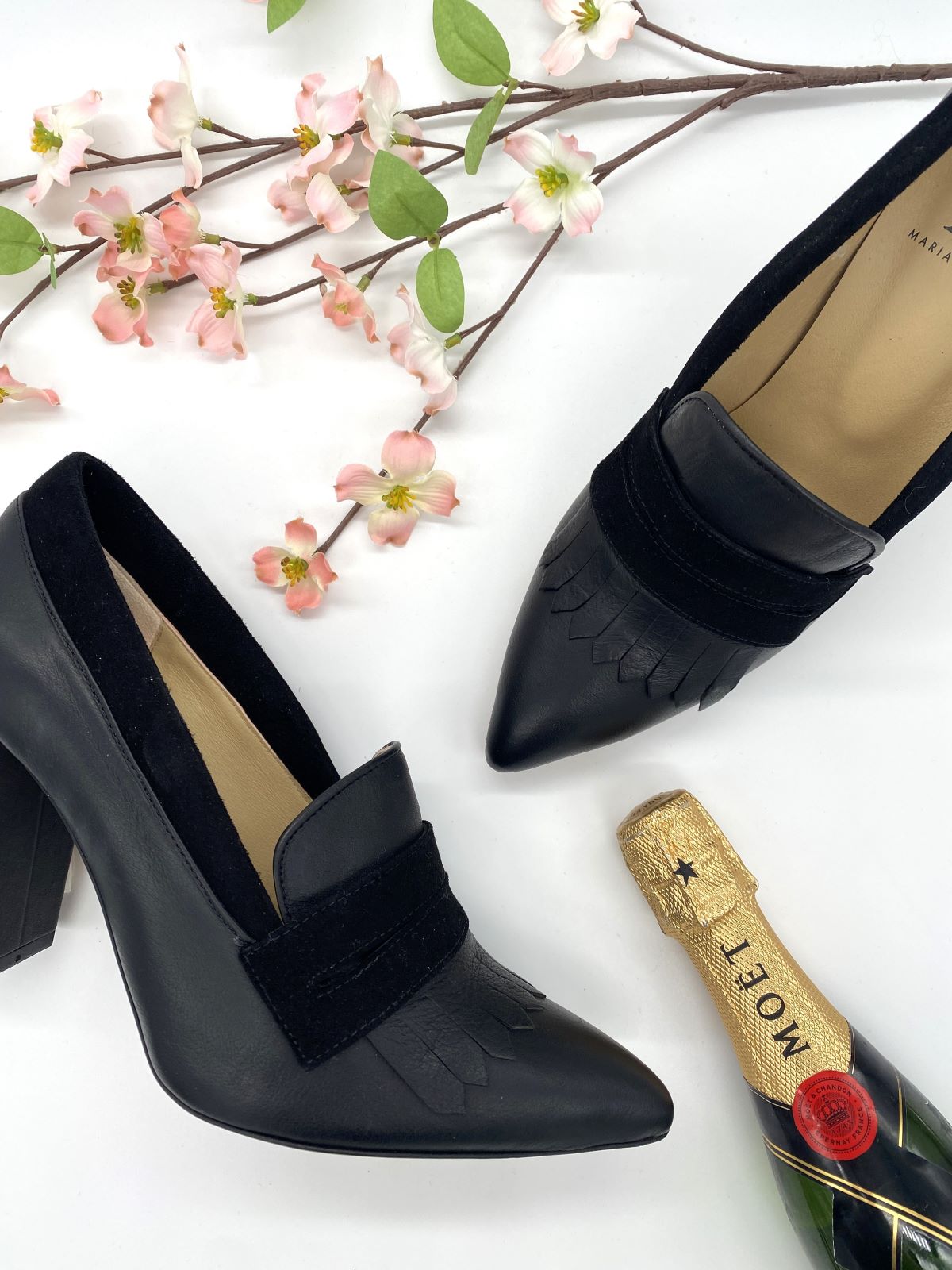 Damenpumps MaYa in schwarz mit mini Flasche Moet und Blumen im Hintergrund